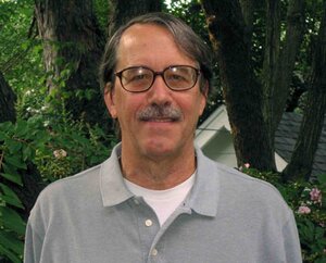 This is a photo of Douglas Barnes. He is wearing a grey polo shirt and standing in front of green foliage.